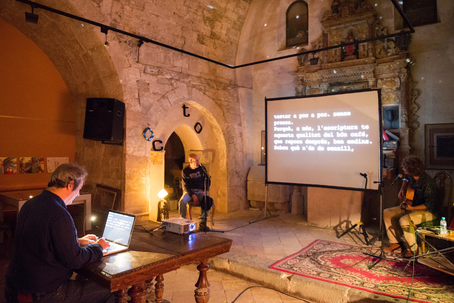 Concierto de Escritura en Cervera: un escritor al teclado, un narrador y un guitarrista, con el texto proyectado sobre la pared de piedra